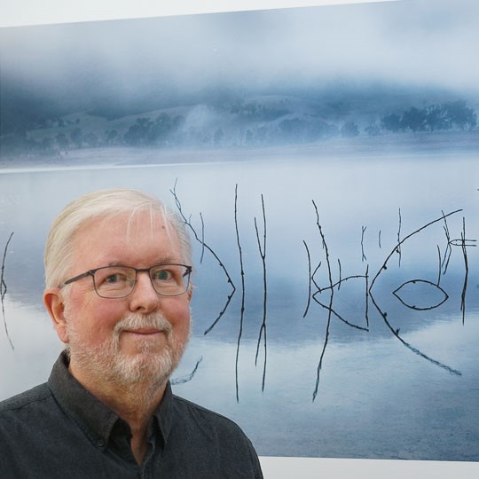 Artist Rene Vogelzang stands in front of a photgraph he has taken. Head and shoulder shot, artist has short grey hair, glowing skin, glasses and a smile on his face. The photo behind him shows a silver looking body of water like a lake with thin sticks protruding out of the water creating circle like pattern 